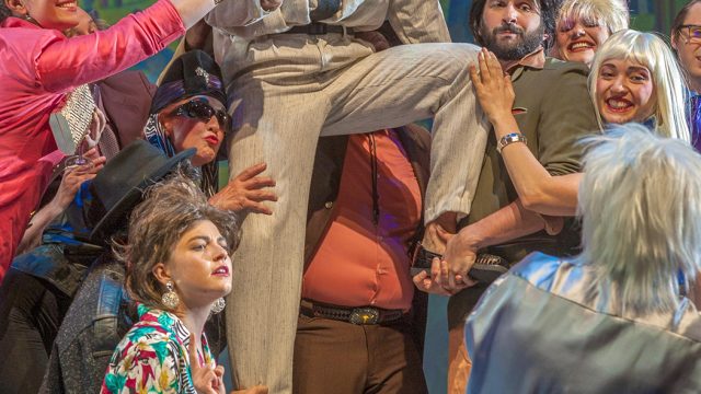 Guests at an art exhibition hoist George (John Bambery*) in the air as they pose for a photograph.