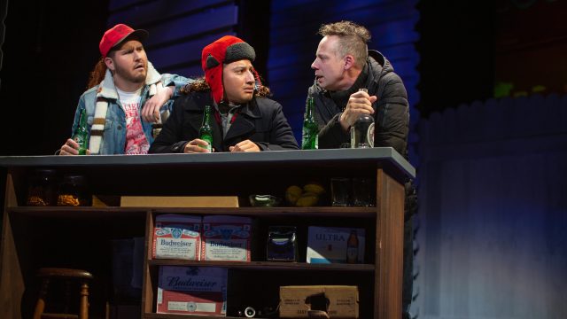 Phil Connors (Ryan Drummond*) gets a drink at the local bar with Gus (Scott Taylor-Cole) and Ralph (Jorge Luis Diaz) after reliving the same day over and over again.