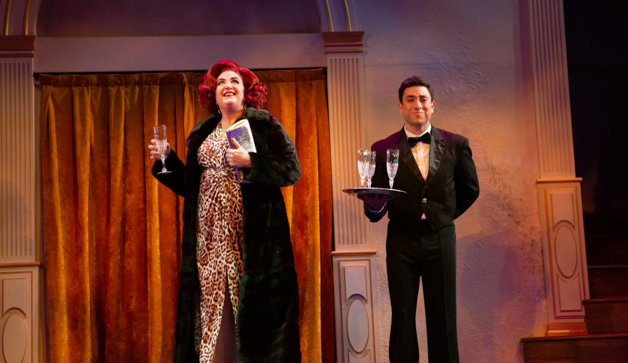 Carlotta Campion (Cindy Goldfield*, left) is greeted by Kevin (Anthony Maglio, right) with a glass of champagne.