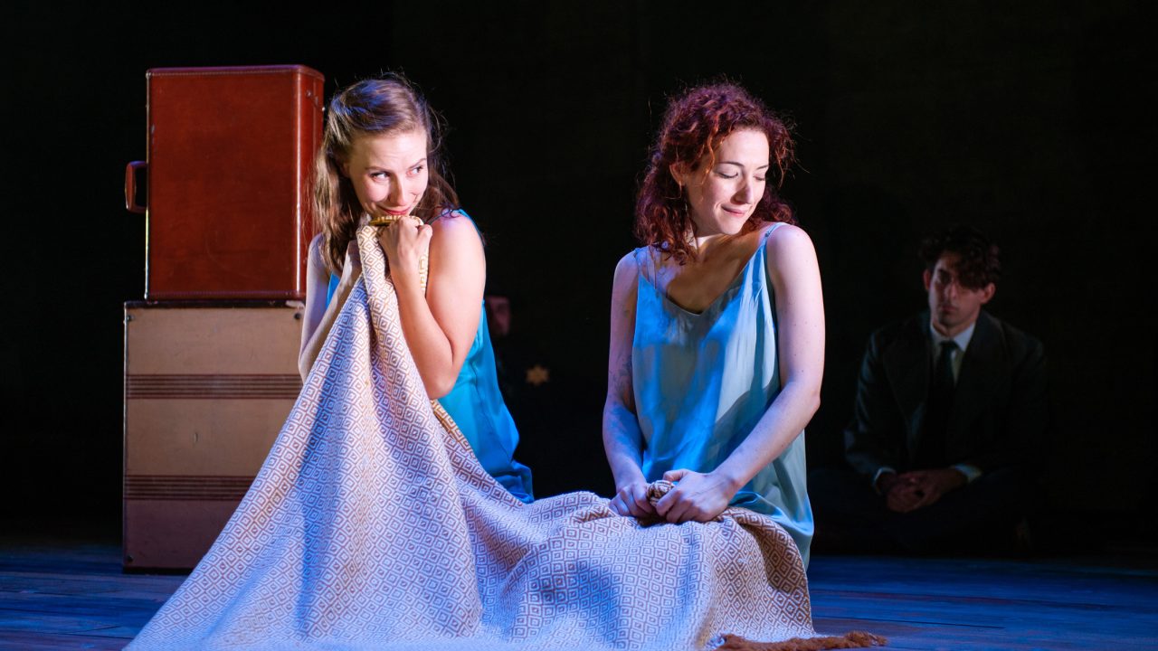The troupe performs Act Two of 'The God of Vengeance' in an attic they transformed into a stage: Manke, played by Halina (Rivka Borek*, left) gets close to Rifkele, played by Chana (Malka Wallick*, right). Troupe member Avram (Billy Cohen*) watches them perform. Production photography by Jessica Palopoli for 'Indecent' by Paula Vogel at San Francisco Playhouse.