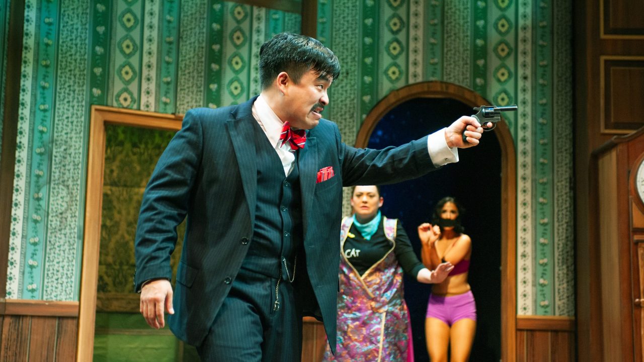 Chris (Phil Wong) wields a gun as performers (Renee Rogoff and Erin Rose Solorio) watch in "The Play That Goes Wrong," presented by San Francisco Playhouse September 21 - November 9. "The Play That Goes Wrong" production photo.